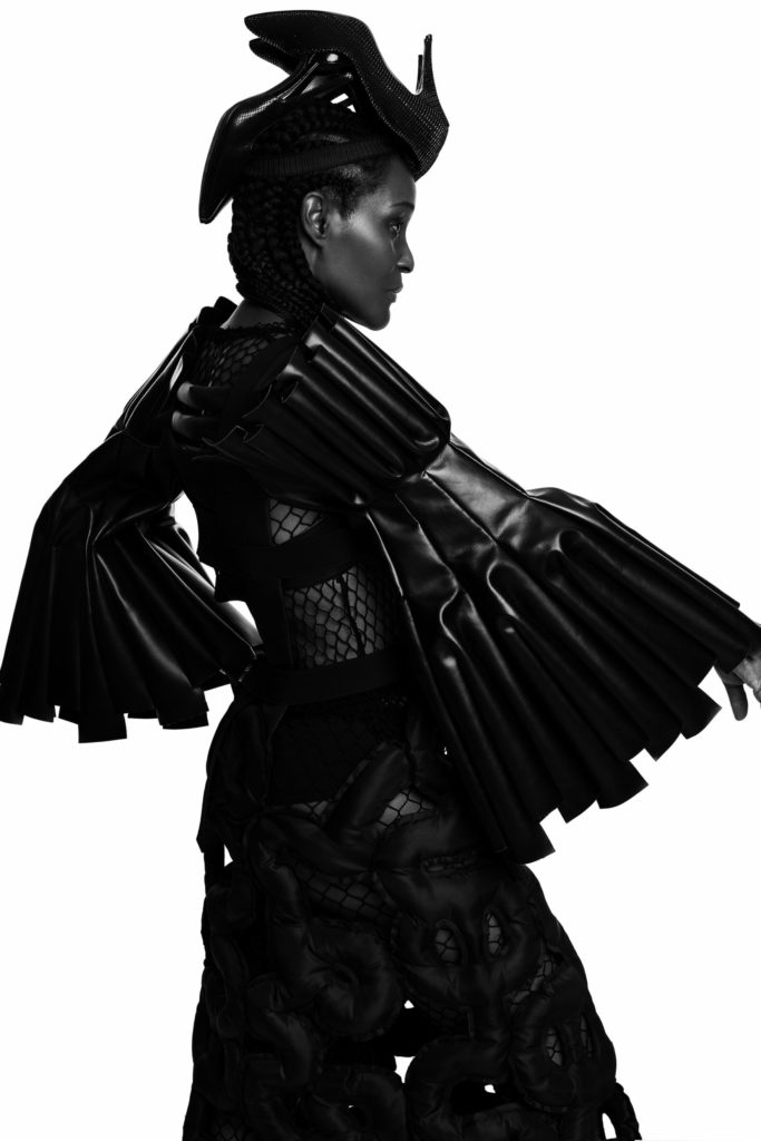 She wears a hat by WILLIE COLE x COMME DES GARÇONS (FW21), and two dresses and an arm protector by COMME DES GARÇONS (SS14).